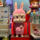 The Monster Mega Labubu Sculpture - 100cm by Giant Sculptures is a pink figure with sharp teeth, upright ears, a skull chest accessory, and a mischievous smile, displayed among colorful toys and figures.