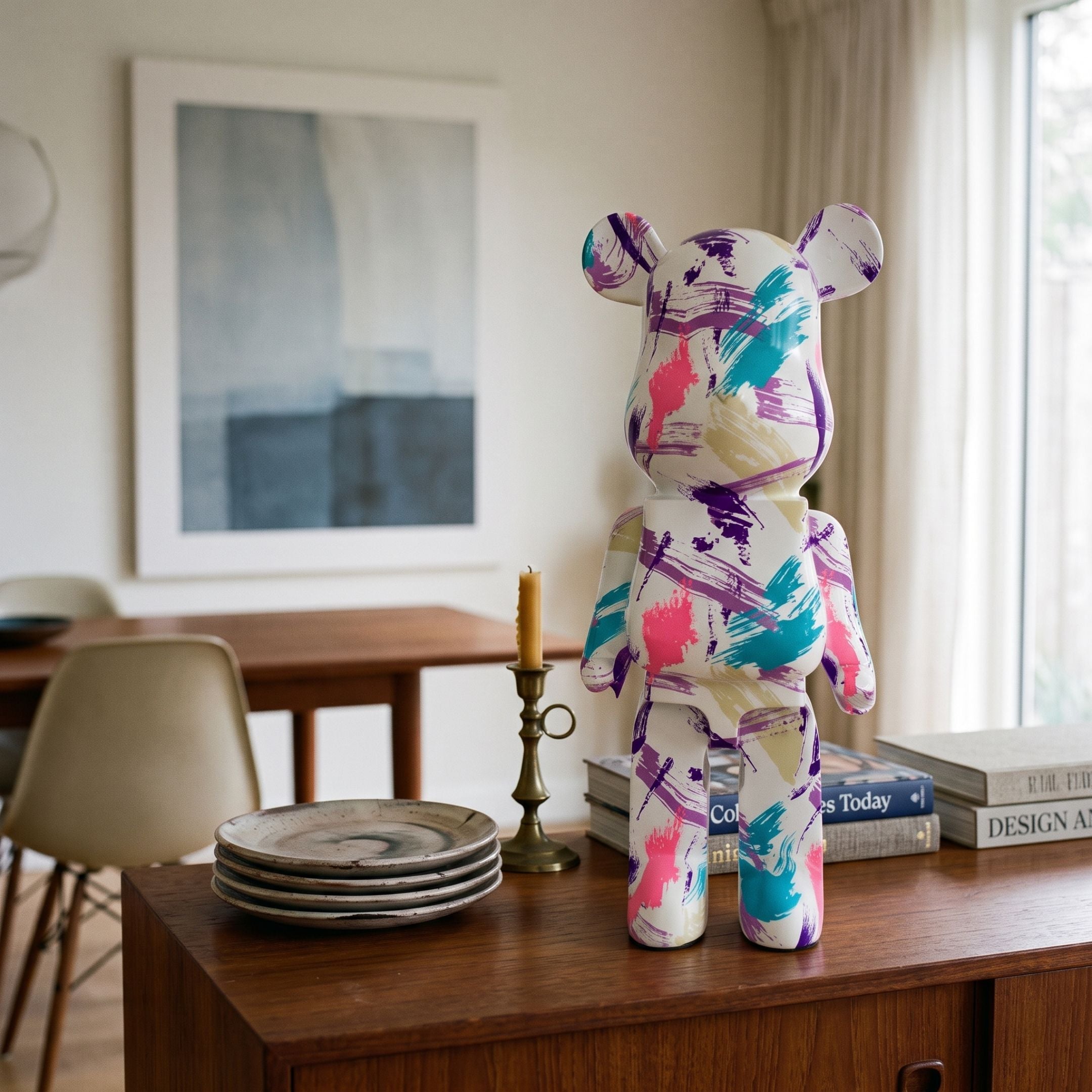 Muse Metro Bear Sculpture - 50cm by Giant Sculptures, hand-painted in purple, teal, and pink brushstrokes on white, displayed on a wooden sideboard beside books and a candlestick.