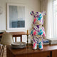 Muse Metro Bear Sculpture - 50cm by Giant Sculptures, hand-painted in purple, teal, and pink brushstrokes on white, displayed on a wooden sideboard beside books and a candlestick.