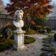 The Apollo Marble Bust Sculpture - 50cm by Giant Sculptures stands on a pedestal in a serene Japanese-style garden, surrounded by autumn foliage, mossy stones, and red and orange leaves scattered along a stone path.