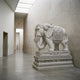 Caparisoned Elephant Grey Granite Statue - 260cm, carved in profile with ornate headpiece and draped panels, displayed on a tiered pedestal in a stone-walled gallery corridor.