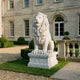 Seated Lion With a Calm Face Marble Statue - 215cm, white marble sculpture on a moulded pedestal, displayed on a stone terrace in front of a manor house.