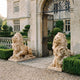 Couchant Lion With Head Raised Marble Statue - 125cm, honey-amber pair flanking a wrought-iron gate at a stone manor entrance, mounted on rectangular slabs.