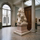 Seated Lion With a Scrolled Cartouche Marble Statue - 210cm displayed on a pedestal in a gallery hall with columns, terrazzo flooring, and an archway view.
