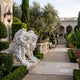 Pair of white marble Crouching Lion With a Spiked Mane Statues, 120cm, flanking a garden pathway beside boxwood hedges and a stone balustrade.