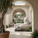 A stone corridor with arches showcases the Majestic Lying Tiger Marble Outdoor Sculpture by Giant Sculptures, set beside a small fountain and surrounded by potted plants, ferns, and lavender beneath a sunlit skylight.