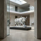 The Powerful Bull White Marble Outdoor Sculpture (150cm) by Giant Sculptures is showcased on a black pedestal in a modern, light-filled gallery with minimalist walls and glass panels, creating an open and airy ambiance.