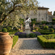 A gravel path leads to the Giant Sculptures Regal Eagle White Marble Outdoor Sculpture (120cm), set among trimmed hedges and purple flowers in front of a stone villa with potted plants and olive trees under a clear sky.