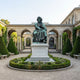 Monumental Classical Seated Male Figure Bronze Sculpture by Giant Sculptures, 400cm verdigris patina figure on a white stone plinth in an arched garden courtyard.