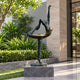 Monumental Contemporary Dancer Bronze Sculpture, 330cm, in a bronze and gold arabesque pose on a stone base, displayed outdoors near landscaped greenery.