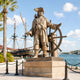 Monumental Traditional Pirate Captain Bronze Sculpture by Giant Sculptures on a stone plinth at a marina, cutlass raised and hand on ship's helm.