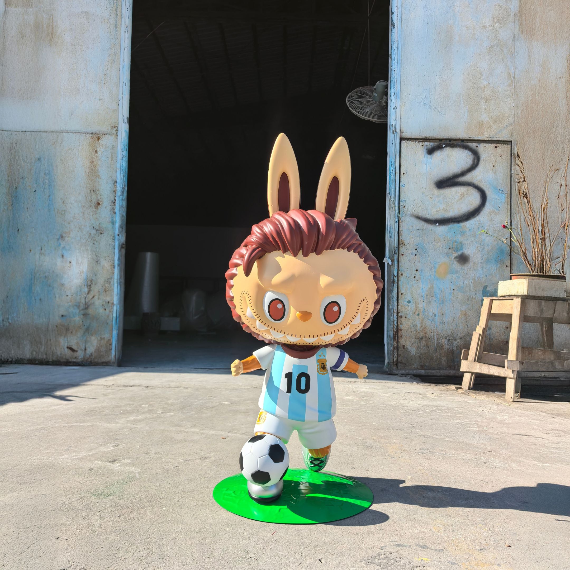 The Giant Sculptures World Cup No.10 Dribble Labubu Sculpture (100cm) features a cartoonish figure with bunny ears in an Argentina #10 soccer jersey, standing on green turf beside a soccer ball, set before an industrial building marked with a spray-painted 3.