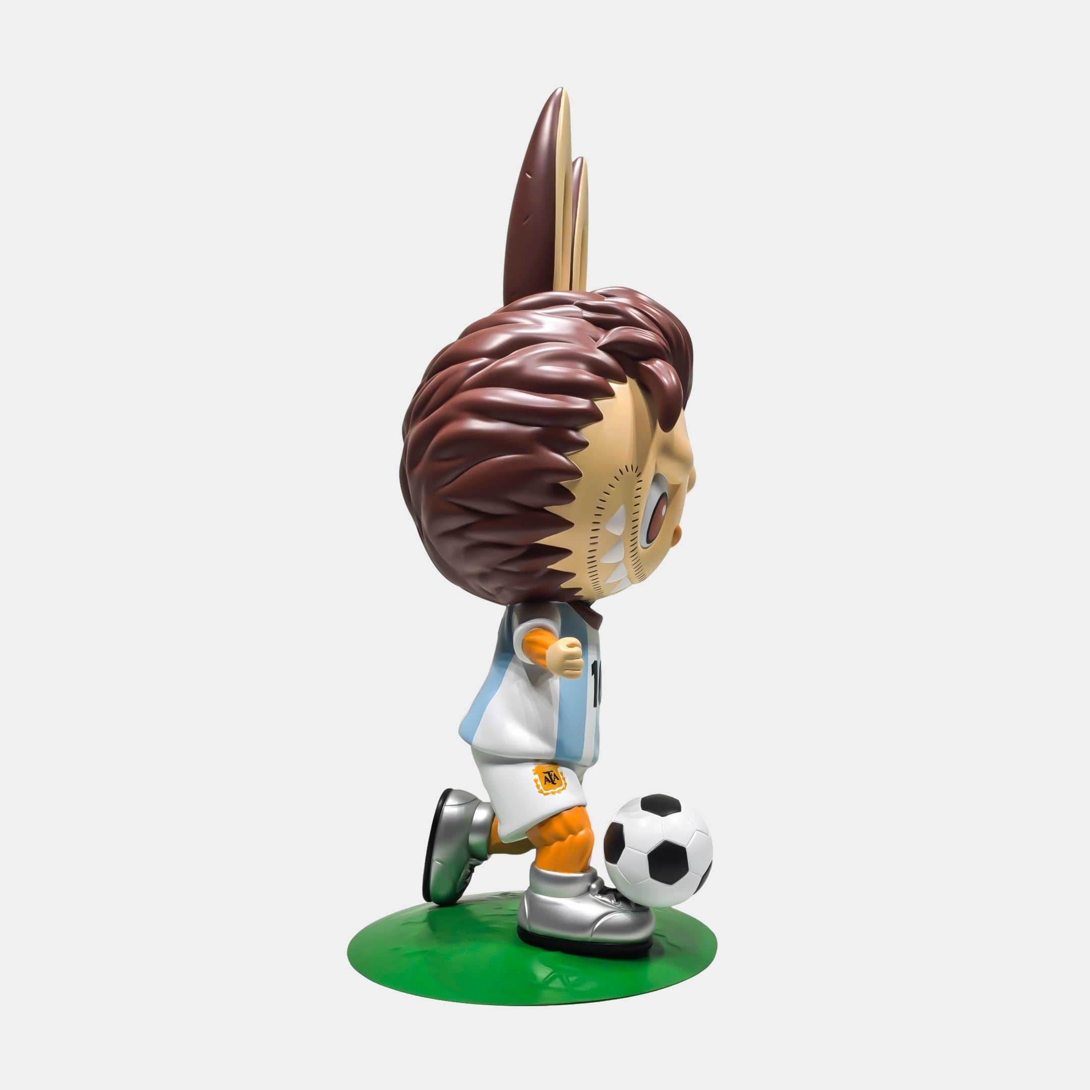 Side view of World Cup No.10 Dribble Labubu Sculpture by Giant Sculptures, 100cm, featuring cartoon-style spiky brown hair, light blue and white soccer uniform, black cleats, on a green base kicking a classic soccer ball.