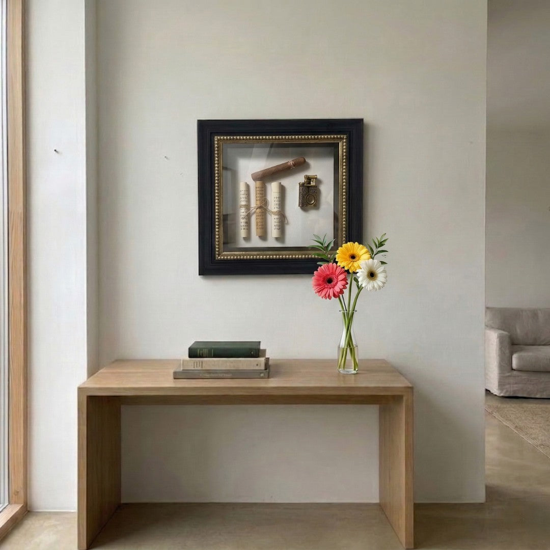 CurioVault Cigar & Sheet Music Framed Wall Art by Giant Sculptures displayed in a hallway above a wooden console table with books and colorful gerbera daisies.