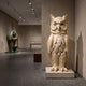 Standing Owl Beige Marble Statue - 150cm displayed on a stone plinth in a gallery, showing carved feather detail and inset amber eyes.