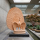 Displaying Peacock Pink Marble Statue, 250cm, shown with wings spread and full tail fan, on a round pedestal in a modern courtyard setting with glass walls and greenery.