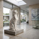 Faceted Cat White Marble Statue - 120cm by Giant Sculptures, sitting upright on a stone base in a modern glass-walled hallway with courtyard views.