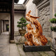 Phoenix on Peony Rockwork Marble Statue - 220cm, carved in brown and cream tones with head turned back and tail sweeping over peony blossoms, set on a dark base in a courtyard.