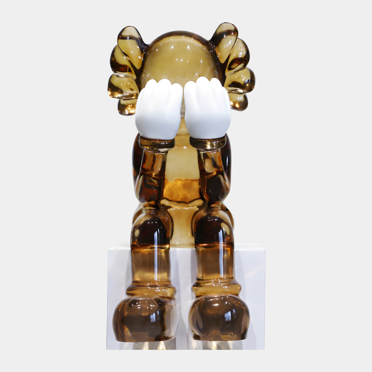 Translucent Amber Iconify Sitting Sculpture, 75cm, resin figure covering face with white hands, seated on white pedestal against plain background.