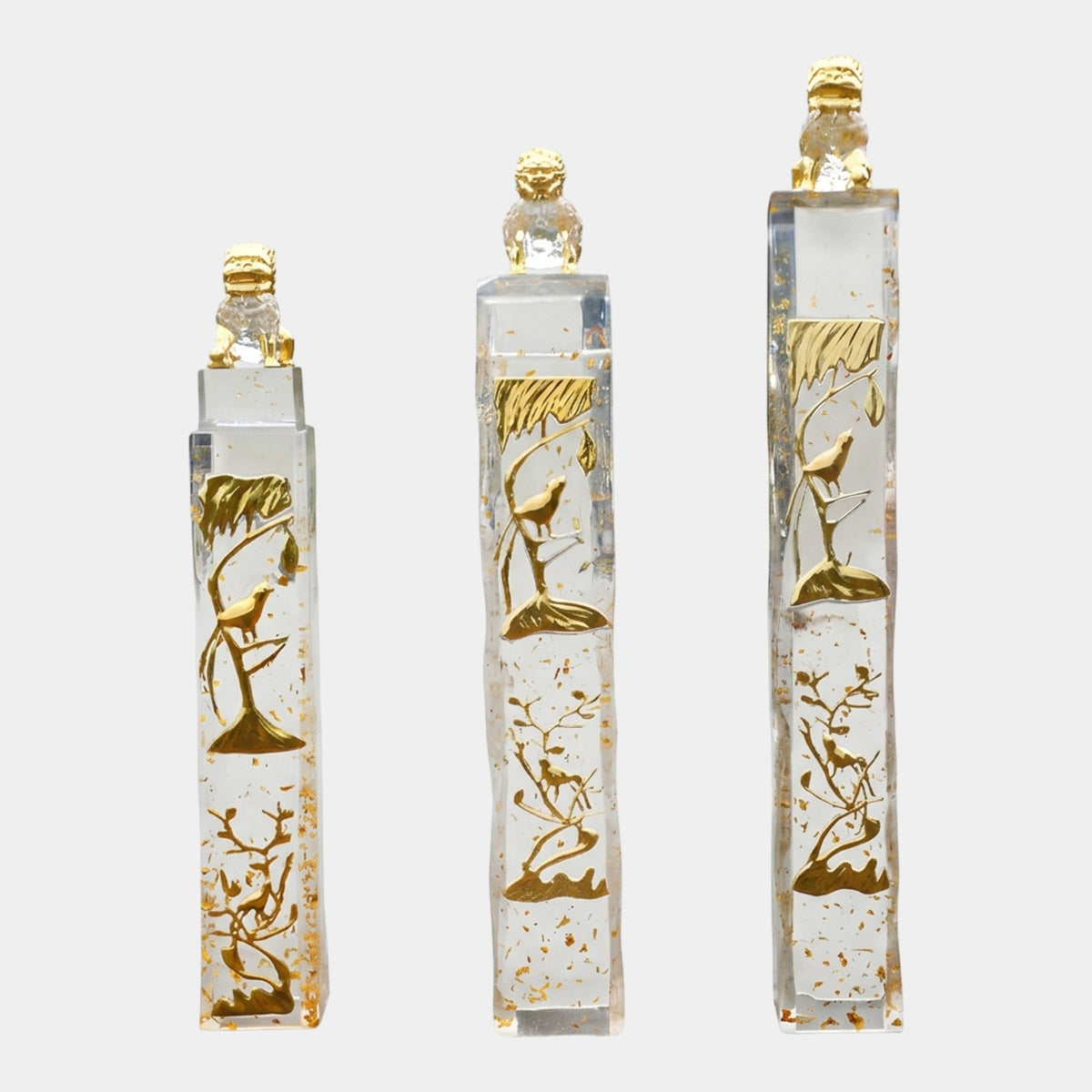Three clear resin columns by Giant Sculptures in 135/158/200cm heights, each topped with a gilded guardian lion and featuring gold tree and bird reliefs, set on a plain white background.
