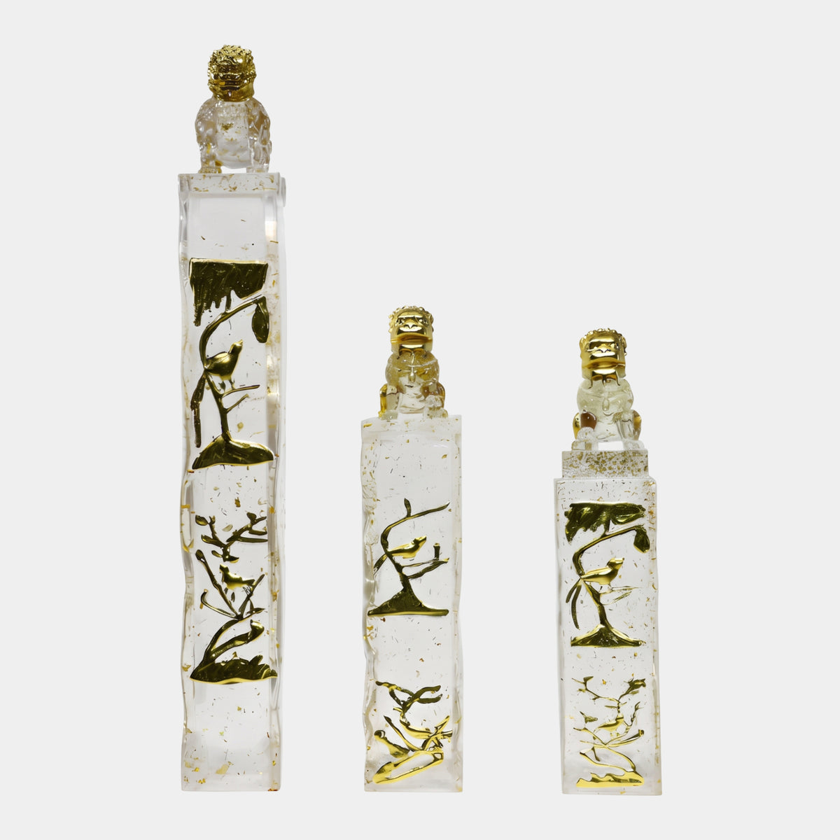 Three clear resin pillars in 89/96/140cm sizes with gold bird-and-branch relief and seated guardian lion caps, gold flecks throughout, on a plain white background.