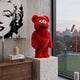 Red Fuzzy Bear Sculpture (70cm) with plush red texture and orange nose displayed on a stone plinth beside a window in a living room with pop art wall decor.