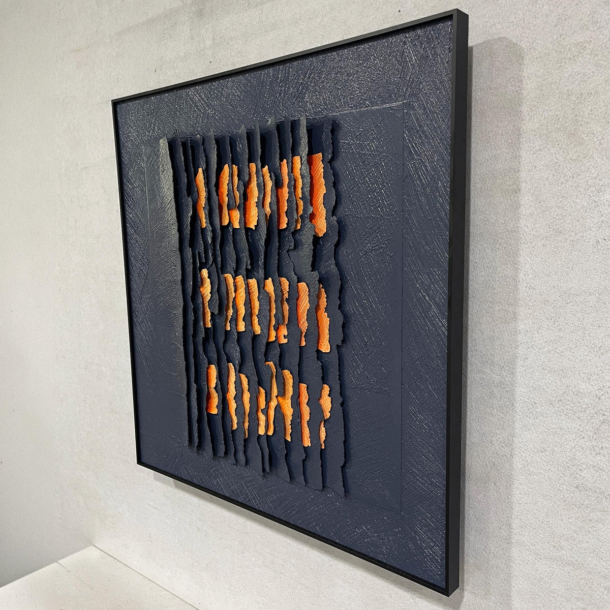 Rivura Navy Layered Sandstone Texture 3D Wall Art in a black frame, showing torn navy strips revealing orange layers, shown at an angle against a light grey wall.