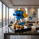 The Giant Sculptures Smurf Couple Hugging Embrace Figure (40cm) stands on a black marble table in a modern living room with large windows showcasing a sunset city skyline. The colorful hand-painted figurine depicts two Smurfs hugging.