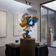 The Smurfette Vanity Mirror Glamour Sculpture - 70cm by Giant Sculptures features a metallic-finished Smurfette holding a vanity mirror, elegantly displayed on a pedestal in a modern gallery with minimalistic and abstract decor.