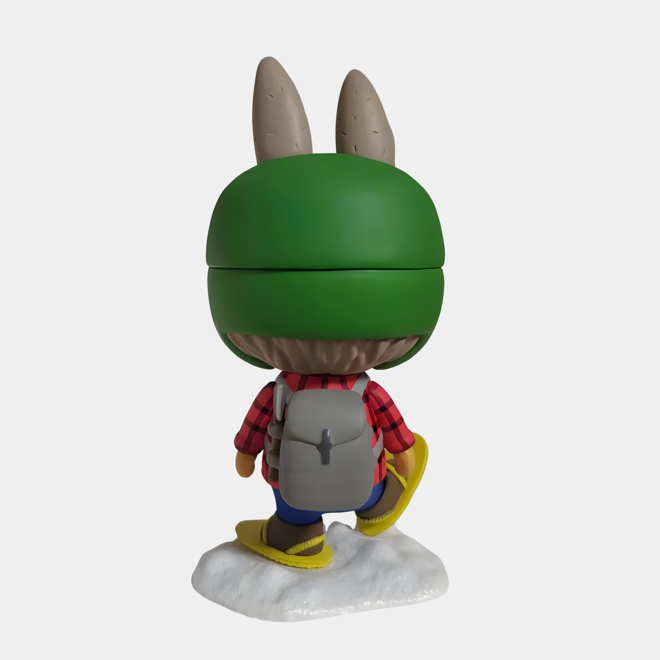 A cartoon-style figure with a green helmet and rabbit ears, red plaid shirt, gray backpack, yellow boots, and blue pants stands on snow—perfect for indoor winter fun with the Giant Sculptures Camp Life Snow Hiker Labubu Sculpture (100cm).