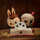 The Spooky Buddies Blade Trick Labubu Sculpture (100cm) by Giant Sculptures features a bunny and a skull with a pink hat behind an open toy book, ideal for unique indoor character displays on wood backgrounds.