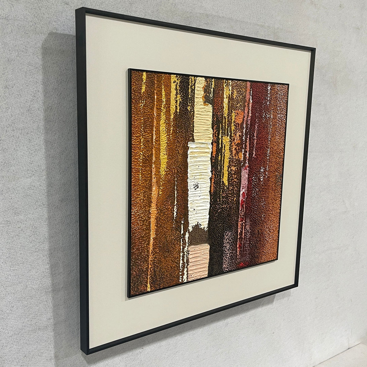 Terralume Amber Rust Abstract Textured Framed Wall Art, side angle view showing vertical brown, gold, and rust-red textured brushstrokes with a central beige stripe, black frame, on a light grey wall.