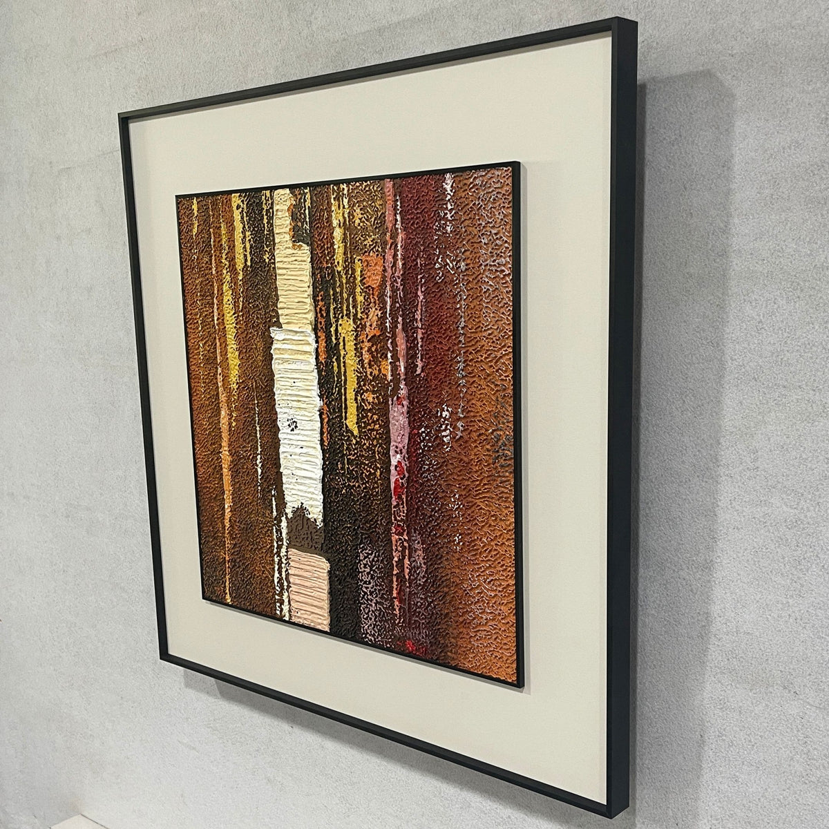 Terralume Amber Rust Abstract Textured Framed Wall Art, side angle view showing textured vertical brushstrokes in brown, gold, and rust tones with a black frame on a grey wall.