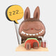 A cute, cartoonish Labubu with rabbit ears naps on sand, smiling with eyes closed, in red shorts and holding a drink—like the 160cm Triangle Chocolate Candy Labubu Sculpture by Giant Sculptures. A “zzz…” speech bubble shows sleep.