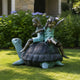 Traditional Children on Tortoise Bronze Sculpture, 140cm, shown outdoors on grass with two children raising their arms atop a spiral-shelled tortoise, set against green shrubs and purple flowers.