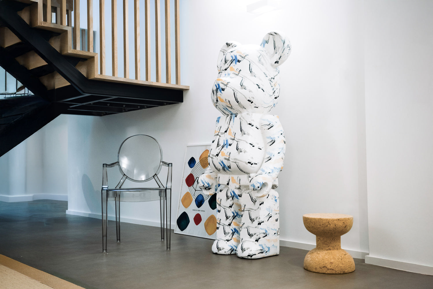 A modern interior with a large, patterned bear sculpture next to a transparent chair and a cork stool. A colorful geometric art piece leans against the wall under a staircase with wooden railings.