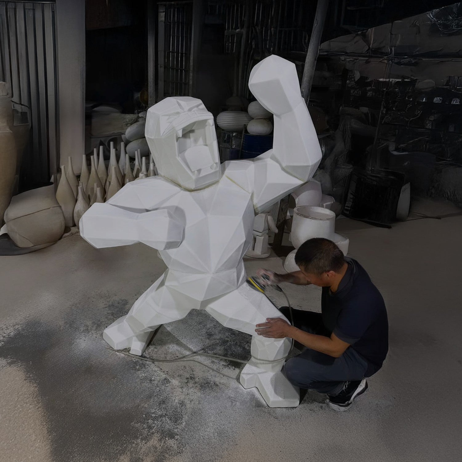 A person works on a large, geometric sculpture of a humanoid figure with a raised fist inside a studio. The sculpture is angular and white, and the studio is filled with various pottery and sculptures.