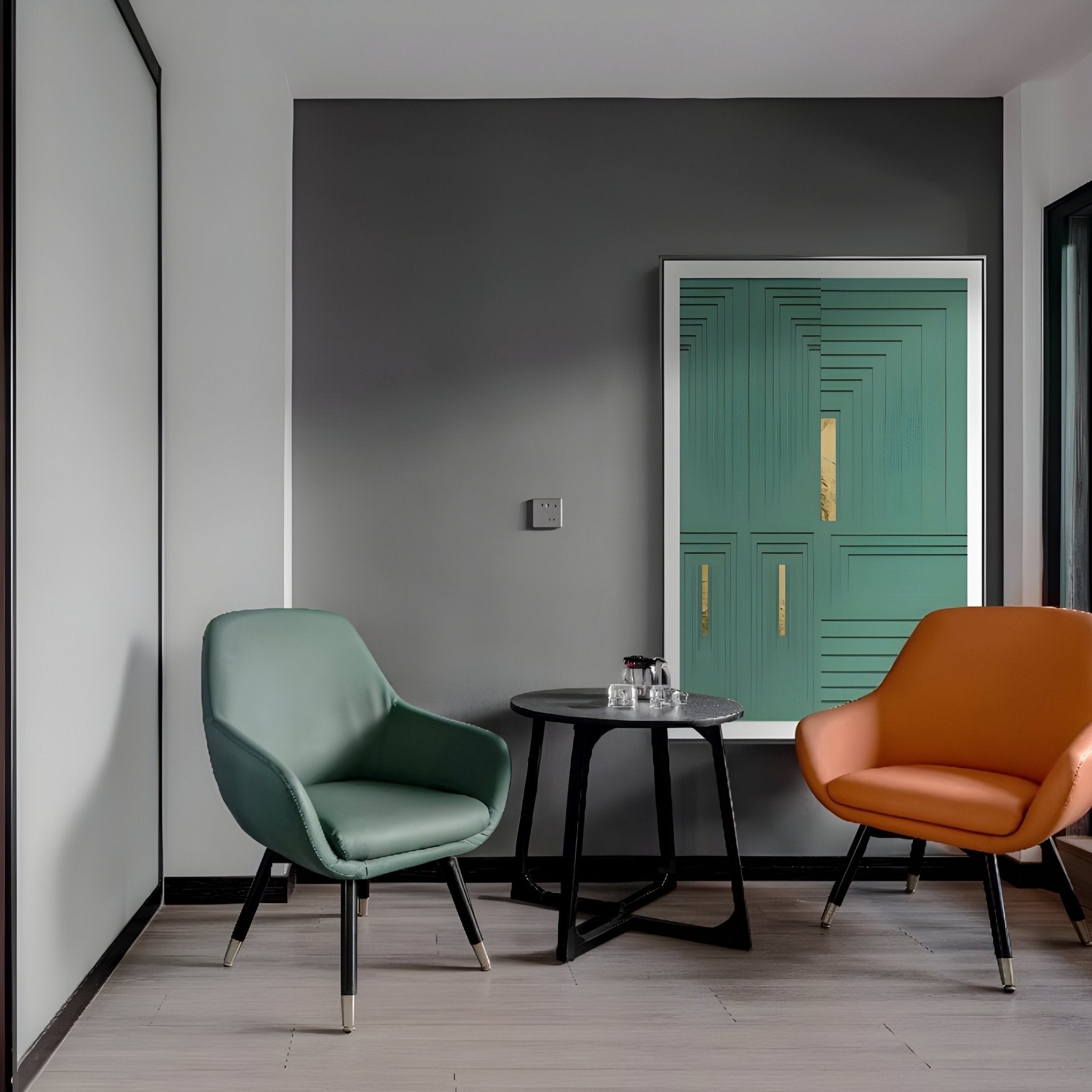 A minimalist room with a gray accent wall includes a green chair, orange chair, and round black table holding two glasses and a jar. On the wall hangs the Giant Sculptures Striato Emerald 3D Wooden Wall Art with Gold Accent, complementing gold decor. The floor is light wood.