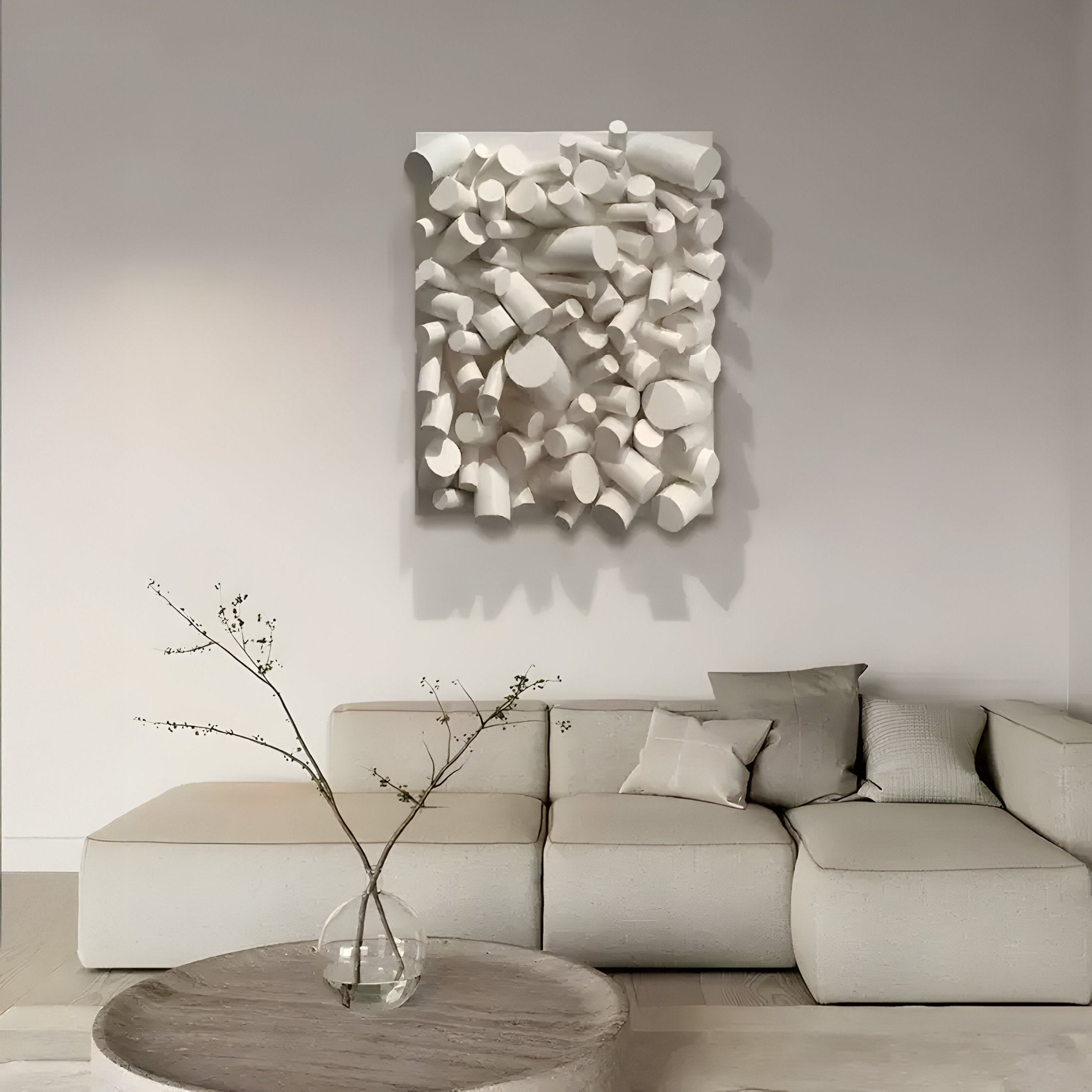 A minimalist living room showcases a light-colored sectional sofa beneath the Forme I Cream Wood Cluster Abstract 3D Wall Art by Giant Sculptures. A round wooden coffee table with a small vase of branches embraces Wabi-Sabi style, adding a natural touch.