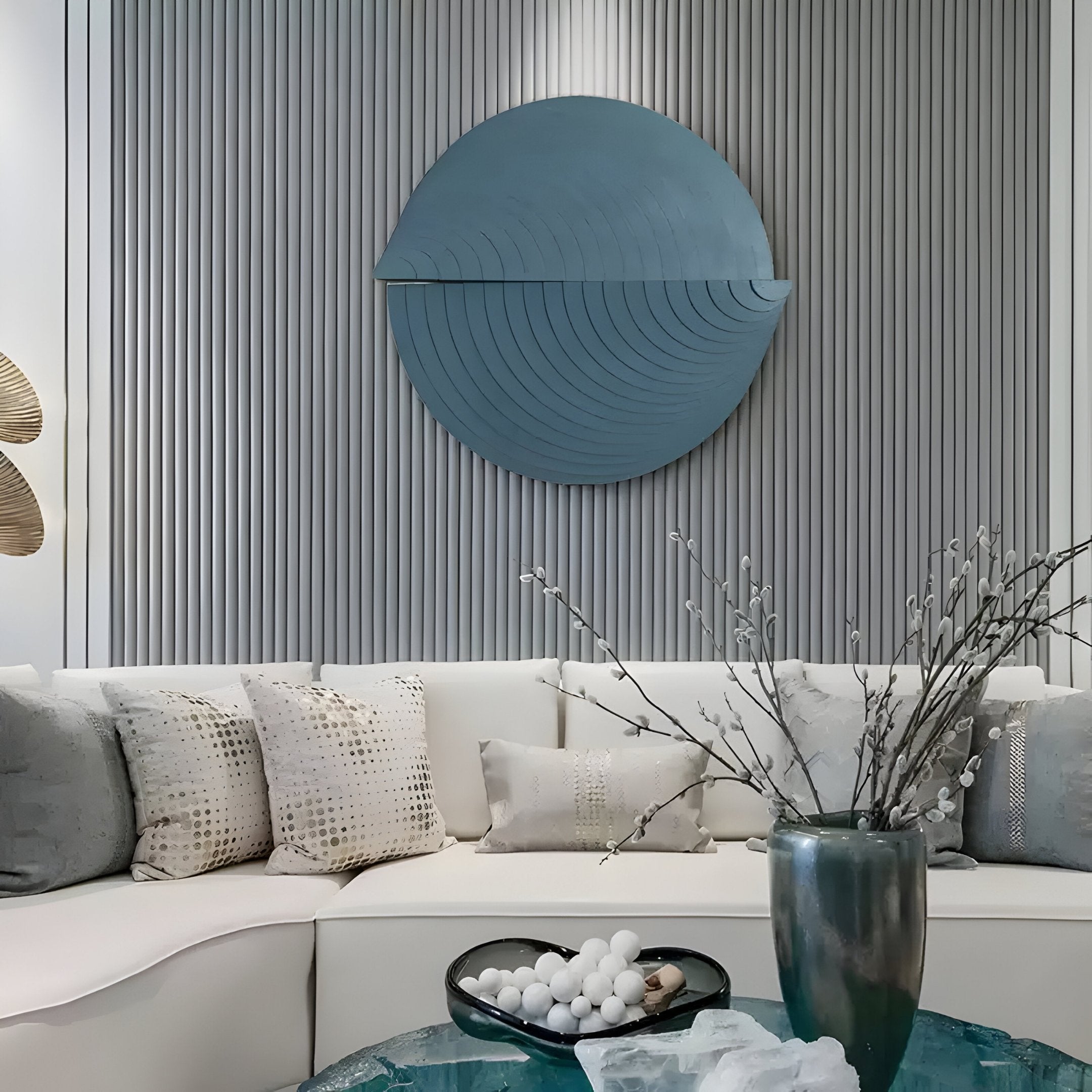 A modern living room features a white sectional sofa with patterned cushions. Orbitals Deep Teal Circular Wood Carved Sandstone 3D Wall Art by Giant Sculptures adorns the striped wall, and a table displays a vase with branches and a bowl of white spheres, enhancing the textured scene.