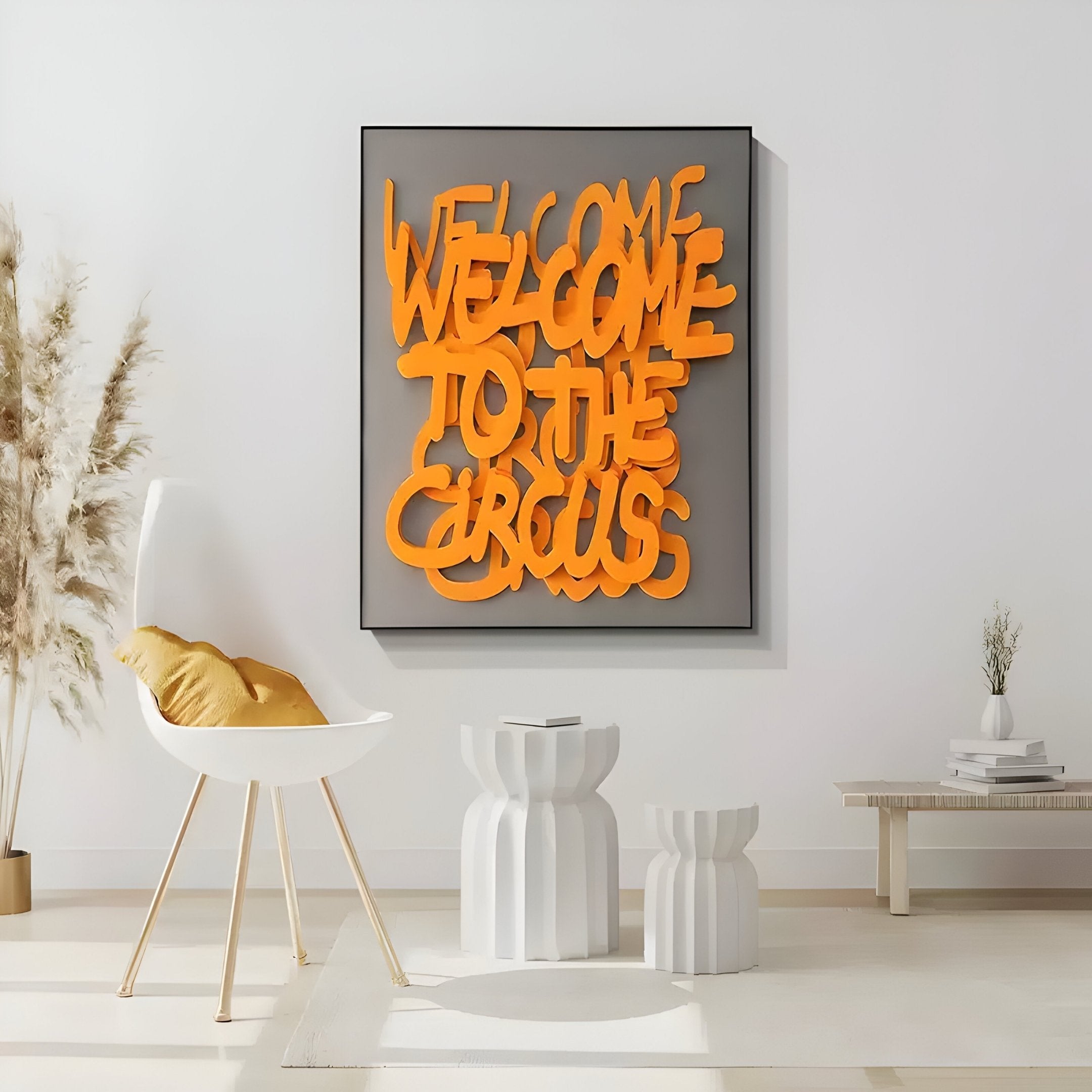 A modern interior features the Urbanic Orange Welcome Script 3D Wooden Graffiti Wall Art by Giant Sculptures, with bold orange text reading Welcome to the Circus. The room includes a white chair with a gold cushion, abstract white tables, beige pampas grass, and a minimalist bench adorned with books.