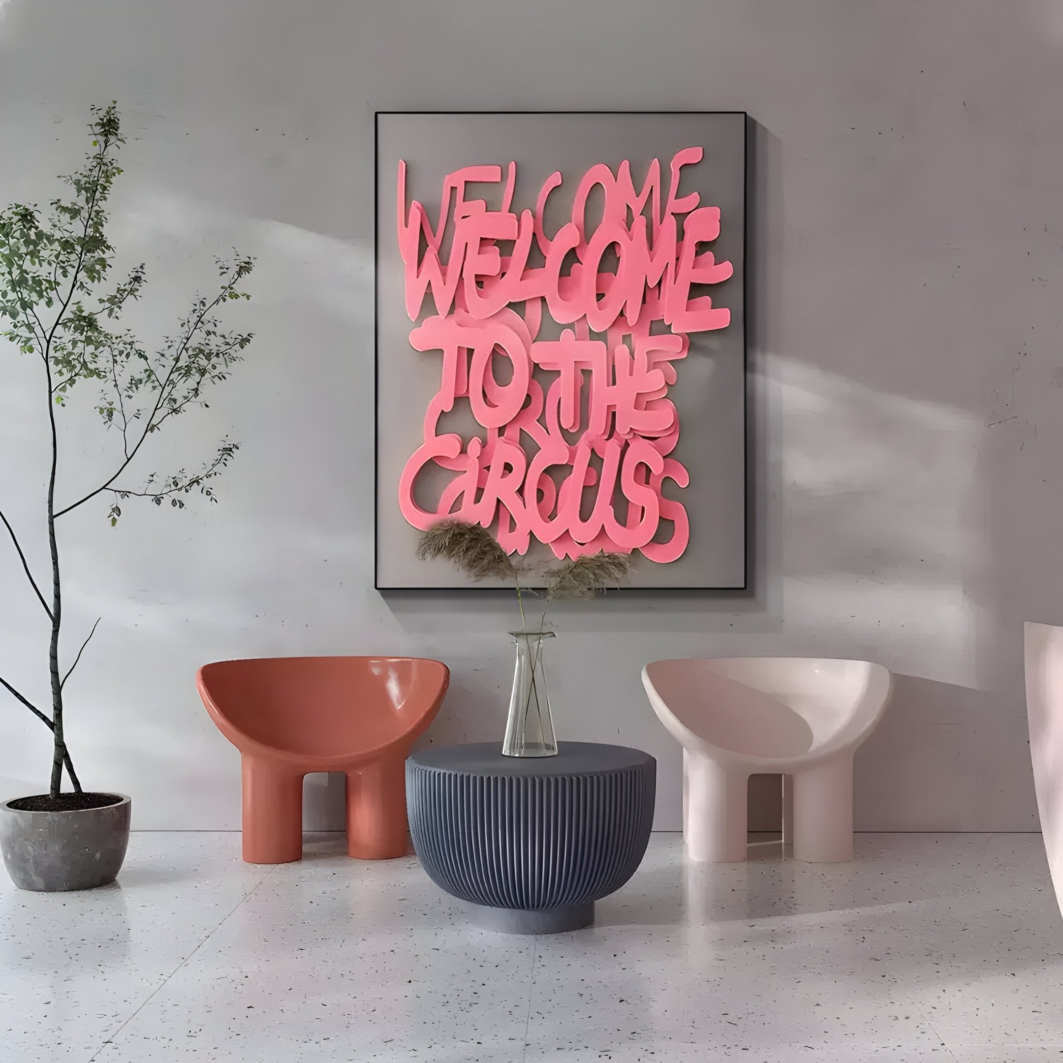 A modern room showcases the Urbanic Pink Welcome Script 3D Wooden Graffiti Wall Art by Giant Sculptures. Below, two distinct chairs—one pink, one orange—flank a gray round table. A potted plant complements this contemporary decor.