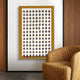 The Artiora Gold Edge & Wood Block Modular Grid 3D Wall Art by Giant Sculptures is mounted on a wall with wooden paneling. A quilted, tan armchair sits nearby, complementing the modern art pieces house-shaped cutouts and yellow frame.