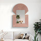 A modern living room boasts a beige sofa with pillows. The Aurellis Pink Arch & Square Geometric Mirror Frame Wall Art by Giant Sculptures hangs above, reflecting a lamp and plant. Potted plants and dried flowers enhance the decor, creating an inviting space.