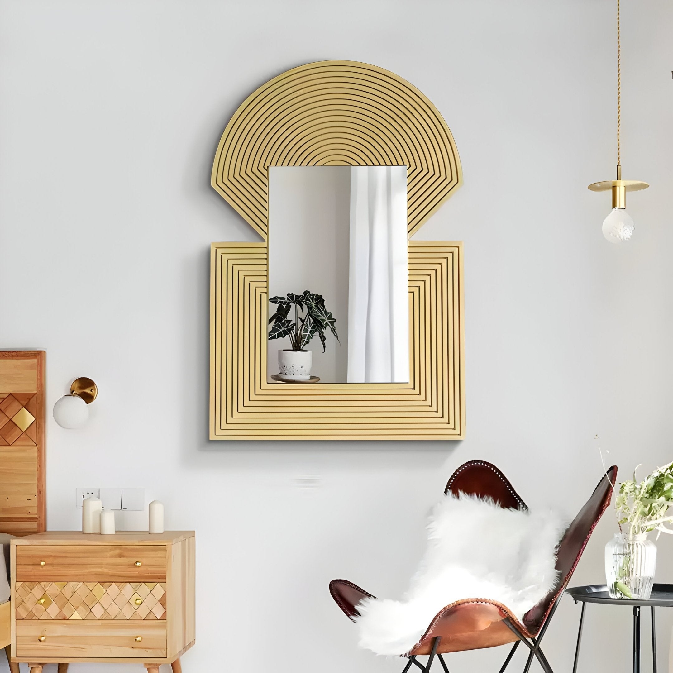 In a modern living room, the Aurellis Gold Arch & Square Geometric Mirror Frame Wall Art by Giant Sculptures reflects a potted plant. Below, a wooden cabinet with candles complements a cozy brown leather chair with a white fur throw. A small hanging light adds warmth to the inviting and contemporary space.
.