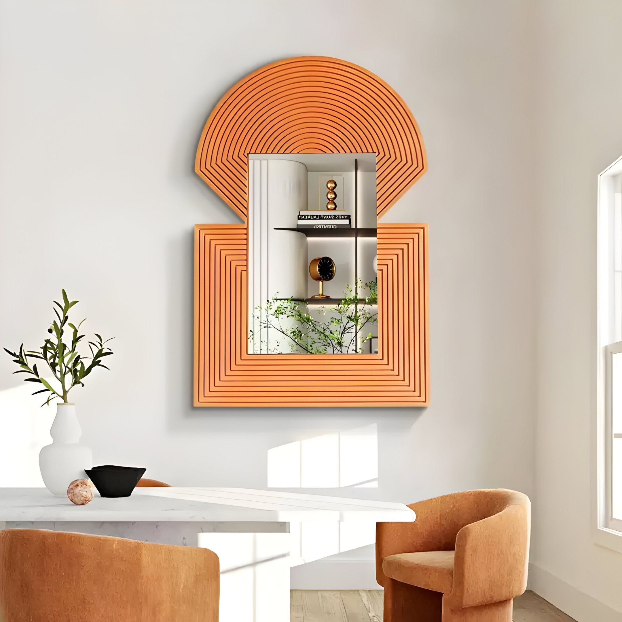 A modern dining room has a round white table with two orange chairs. The wall features the Giant Sculptures Aurellis Terracotta Arch & Square Geometric Mirror Frame Wall Art, reflecting a shelf with decor and a plant. Natural light pours in from the window on the right.