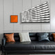 A modern living room has a black leather sofa with black, white, and orange pillows. The wall displays the Illustra Minimalist BOOM Text 3D Wooden Wall Art by Giant Sculptures in square orange typography and features a fan-shaped light emitting a patterned glow.