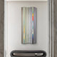 The Illumina Chrome Colourful Vertical Wooden Hand-Painted 3D Wall Art by Giant Sculptures, featuring gray, blue, and orange stripes, adorns a white wall above a minimalist gray upholstered bench, with marble accents enhancing its contemporary flair.