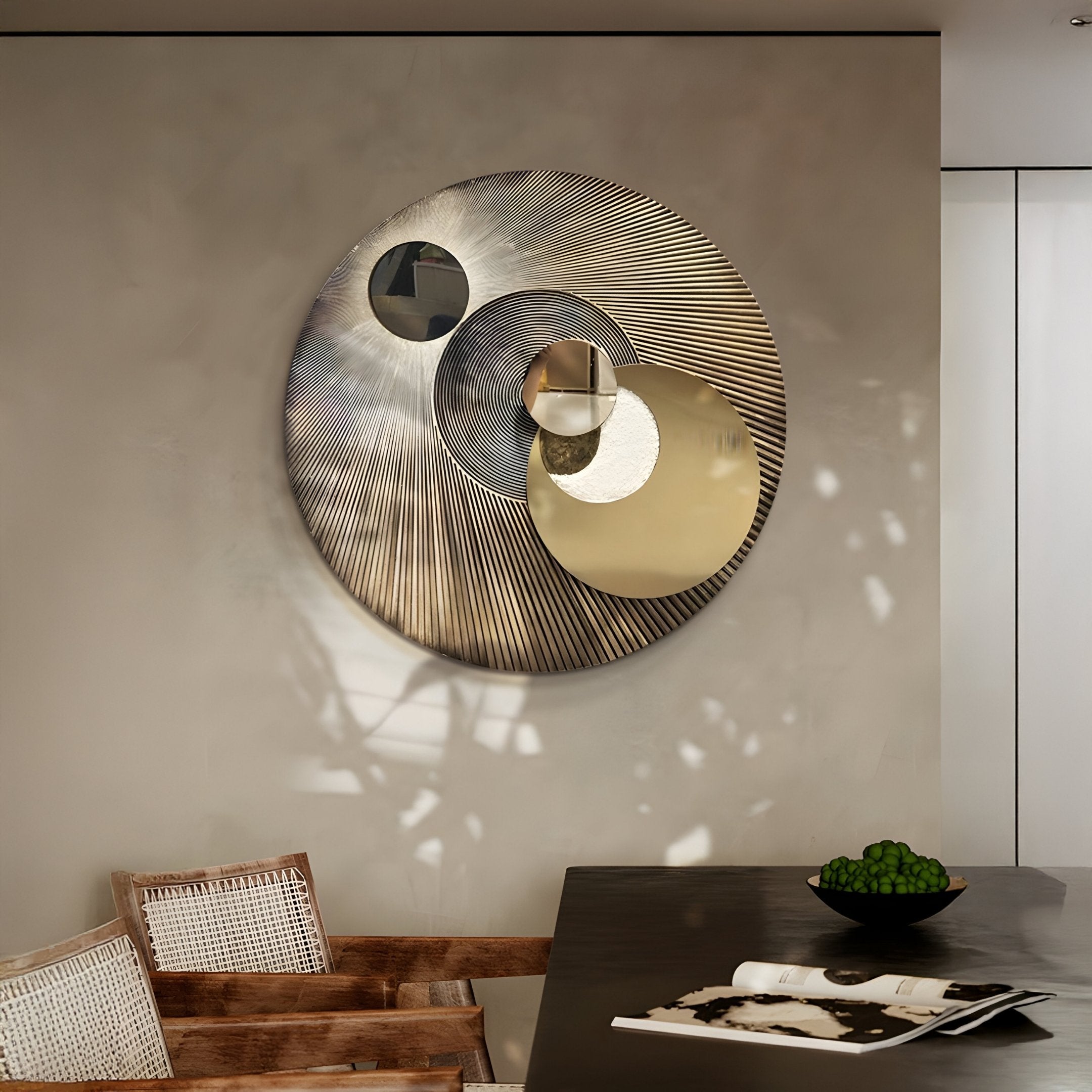 A modern interior features the Orbitra Gold Reflective Circular 3D Wall Art by Giant Sculptures, casting shadows on a beige wall. Below, a dark table holds green apples and a magazine. Two wicker chairs to the left complete the circular design theme.