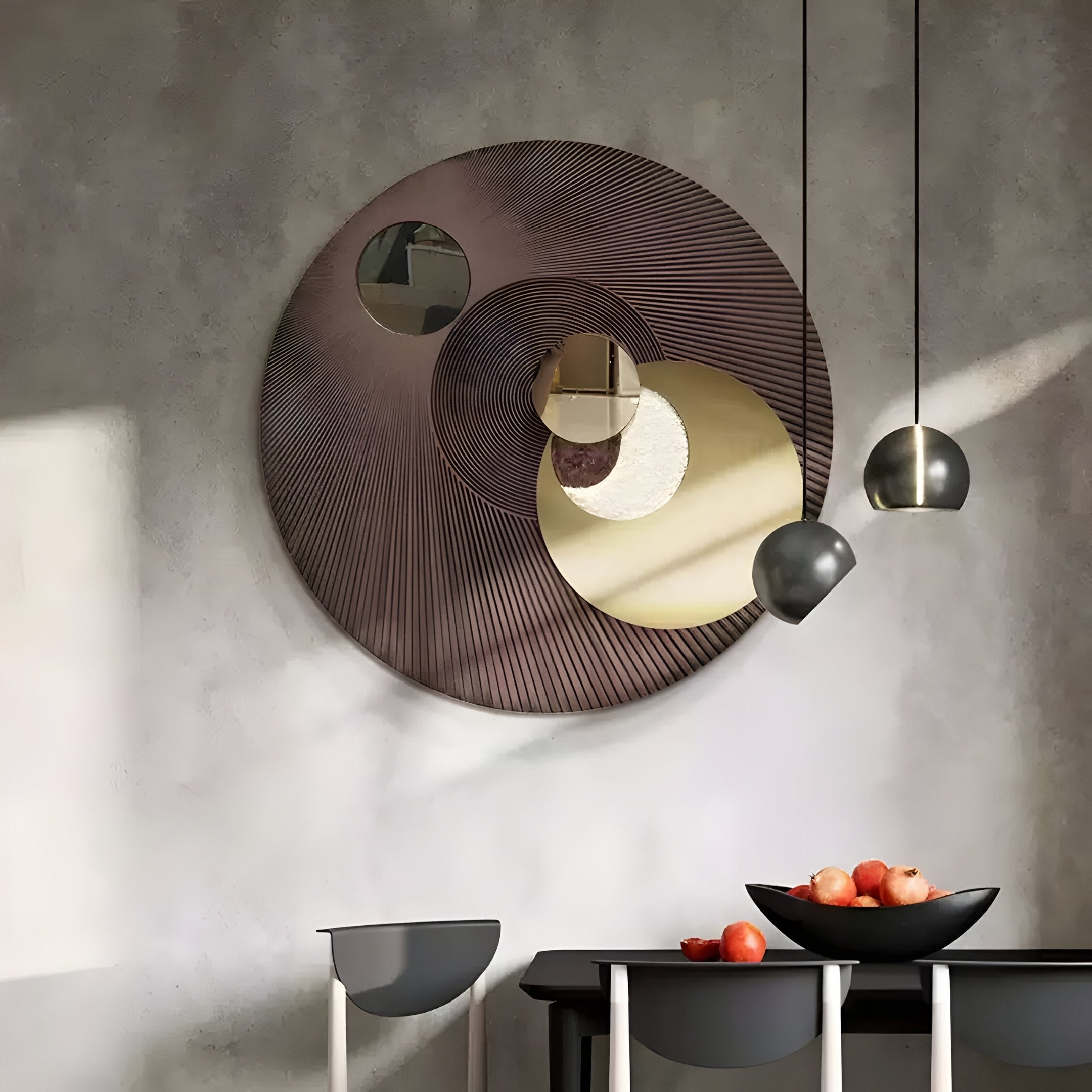 A modern interior features the stunning Orbitra Bronze Reflective Circular 3D Wall Art by Giant Sculptures, showcasing concentric circles. Below, a table holds a black bowl of apples beneath two pendant lights. A chair to the left adds balance to the space.